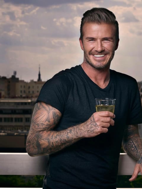 Plan moyen de David Beckham debout sur un balcon avec en toile de fond le paysage urbain de Barcelone, il a une contre-dépouille texturée élégante avec un côté délavé et des cheveux balayés vers l'arrière, des tatouages ​​sur les manches complètes sur les deux bras, porte un t-shirt noir à col en V, tient un verre avec une boisson jaune et sourit largement à la caméra.