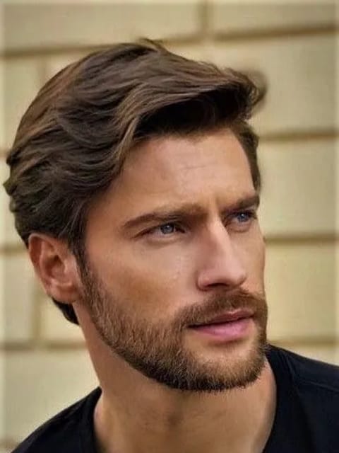 Un portrait en gros plan d'un homme aux cheveux bruns épais et coiffés avec des vagues douces et de subtils reflets marron clair. Il porte un haut noir à col rond, placé devant un mur carrelé en pierre beige clair, regardant hors caméra avec une expression calme. Modèle pour l'échange de visage d'image.