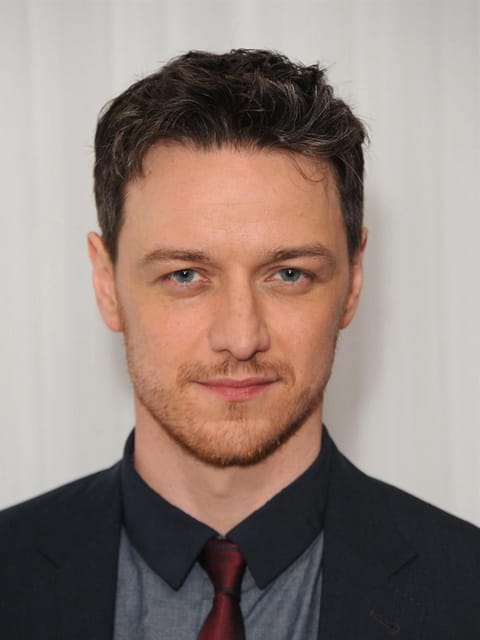 Portrait de James McAvoy aux cheveux noirs courts et ondulés striés de chaume gris et rouge roux. Il porte un blazer bleu marine foncé sur une chemise à carreaux bleu clair, une cravate rouge foncé et un maillot de corps à col foncé, sur un fond texturé blanc uni.