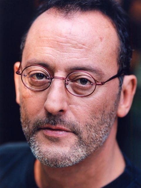 Gros plan de Jean Reno avec ses cheveux noirs courts et dégarnis, ses lunettes rondes cerclées de fil à monture rougeâtre, sa barbe et sa moustache poivre et sel. Il porte un haut bleu foncé à col rond et regarde calmement la caméra.