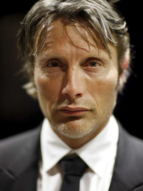 Portrait de Mads Mikkelsen avec des vagues texturées en désordre, dans un costume noir avec une cravate noire, regardant la caméra avec une expression calme.