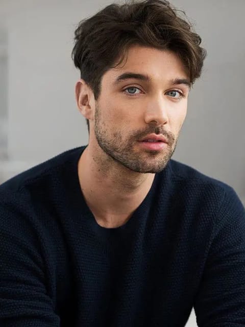 Portrait d'un homme aux cheveux brun foncé épais et ondulés, coiffés dans un look texturé en désordre, dans un pull bleu marine, assis dans une scène d'intérieur avec une expression calme. Modèle pour l'échange de visage d'image.