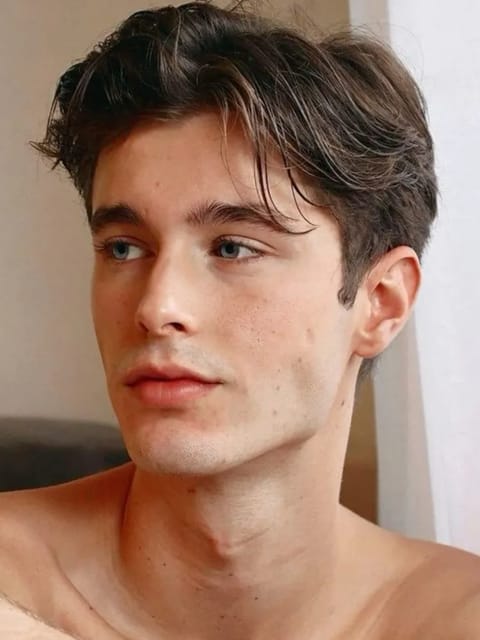 Portrait d'un homme torse nu aux cheveux bruns épais et ondulés, assis dans une scène d'intérieur, regardant hors caméra avec une expression calme. Modèle pour l'échange de visage d'image.