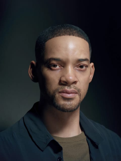Portrait de Will Smith avec ses cheveux courts et foncés et sa barbe claire, il porte une chemise bleu marine à col déboutonné sur un maillot de corps beige à col rond et regarde directement la caméra sur un fond lisse et dégradé gris foncé. Modèle pour l'échange de visage d'image.