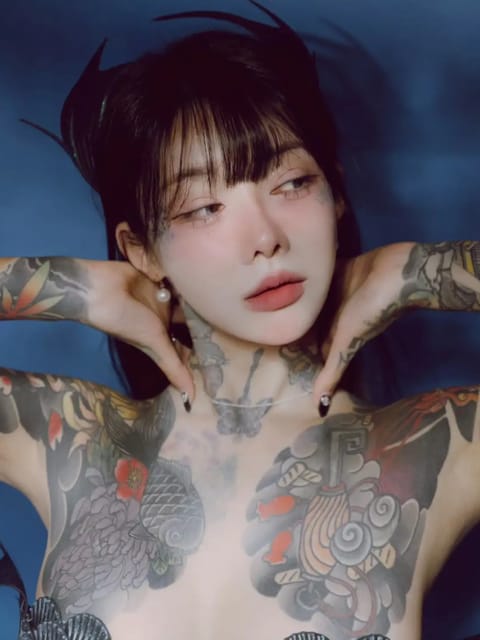 Portrait d'une femme aux cheveux noirs avec une frange émoussée et un tatouage floral d'inspiration japonaise sur la poitrine et l'épaule. Elle pose dans l'eau, les deux mains sur la nuque. Modèle pour l'échange de visage d'image.