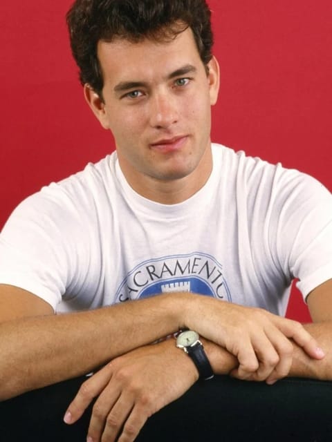 Portrait en studio du jeune Tom Hanks aux cheveux courts, épais et bouclés. Il porte un t-shirt blanc à col rond avec un imprimé graphique bleu, pose les bras croisés sur une surface, a une montre-bracelet au poignet gauche et regarde la caméra sur un fond rouge vif uni. Modèle pour l'échange de visage d'image.
