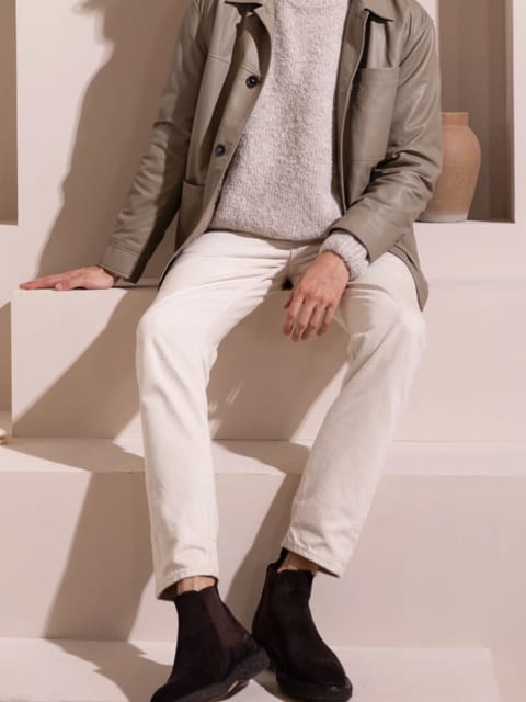 En mann i en grå blazer, lys grå genser og hvite bukser, sitter i et minimalistisk rom, modell for AI-klærbytte.