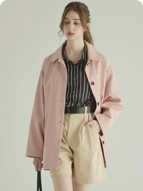 En kvinne i rosa jakke, stripet skjorte og beige shorts, holder en beige veske, i brune støvler, modell for AI-klærbytte.
